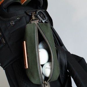 Puede incluir: Una bolsa de golf de lona verde oscuro con cremallera, sujeta a una bolsa de golf negra. La bolsa contiene varias pelotas de golf blancas. Un pequeño detalle de cuero es visible en la bolsa.
