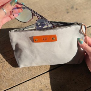 May include: A light gray canvas glasses case with a brown leather tag that has the initials "LK" embossed on it. The case is open, revealing a pair of eyeglasses with a tortoiseshell pattern frame. The case has a zipper closure.
