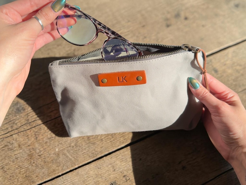 May include: A light gray canvas glasses case with a brown leather tag that has the initials "LK" embossed on it. The case is open, revealing a pair of eyeglasses with a tortoiseshell pattern frame. The case has a zipper closure.
