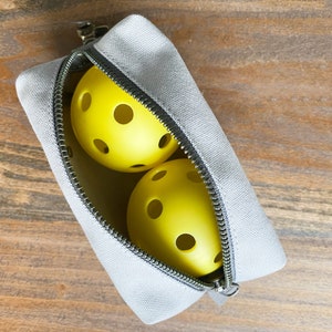 May include: A gray zippered pouch containing two yellow pickleballs. The pickleballs have multiple circular holes. The pouch is open, revealing the balls inside. The background is a wooden surface.