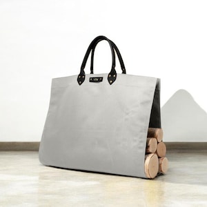 May include: A light grey tote bag with black leather handles and accents. The bag is filled with several pieces of firewood. The bag has a rectangular shape and is sitting on a light-coloured surface.