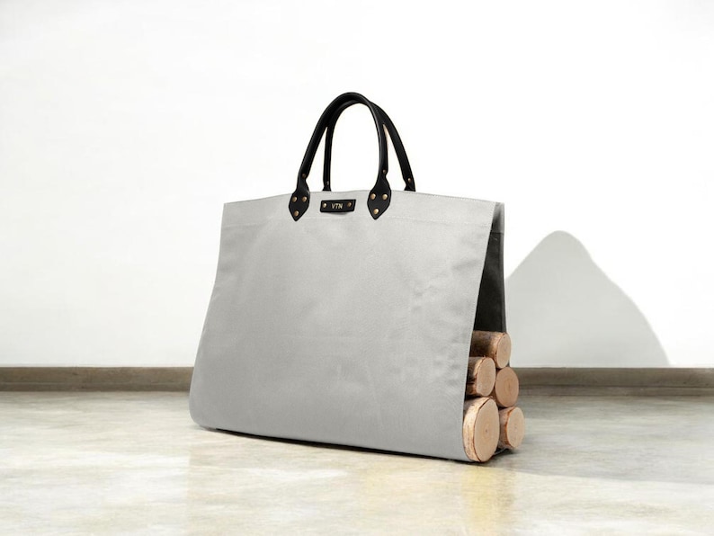 May include: A light grey tote bag with black leather handles and accents. The bag is filled with several pieces of firewood. The bag has a rectangular shape and is sitting on a light-coloured surface.