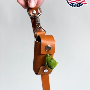 May include: A tan leather waste bag dispenser featuring a silver metal snap closure and a silver swivel clip attached to a matching leather strap. A single green plastic waste bag is visible protruding from the center of the cylindrical holder.