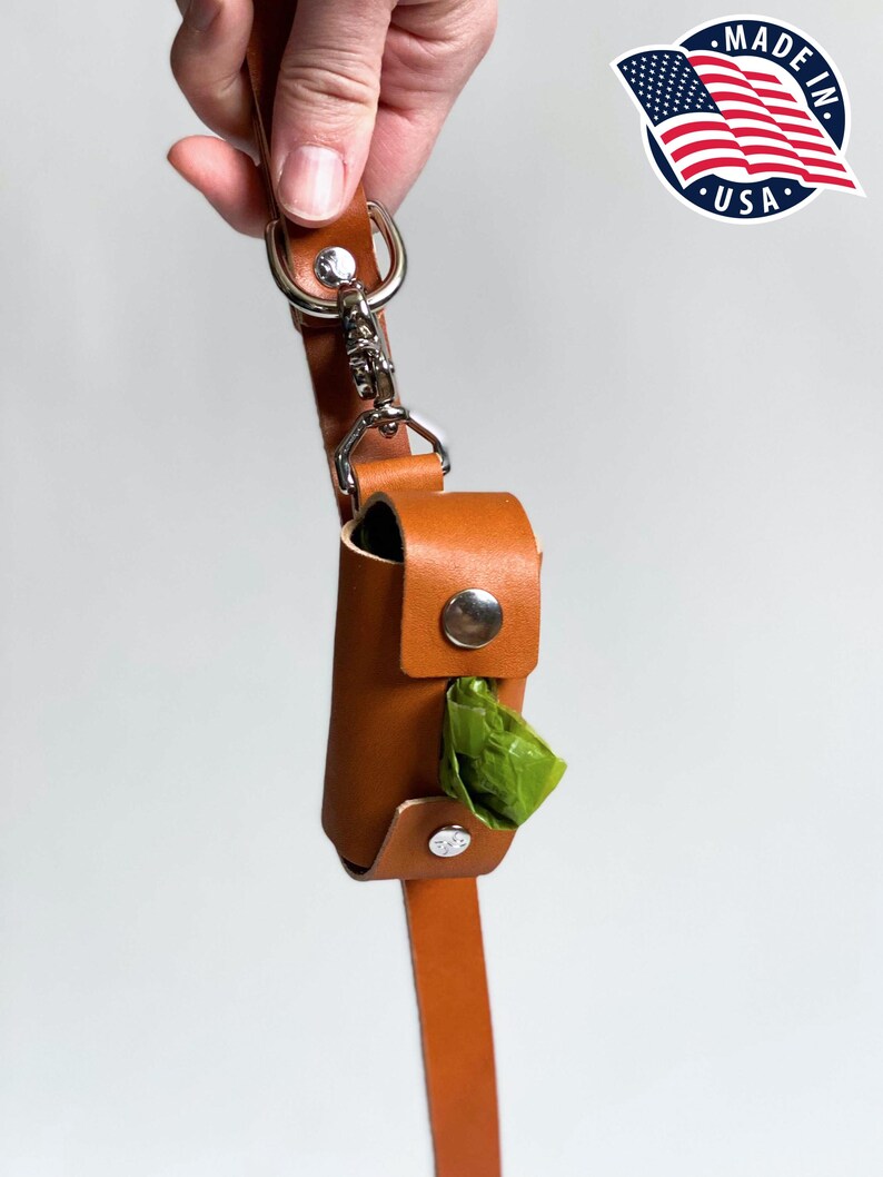 May include: A tan leather waste bag dispenser featuring a silver metal snap closure and a silver swivel clip attached to a matching leather strap. A single green plastic waste bag is visible protruding from the center of the cylindrical holder.