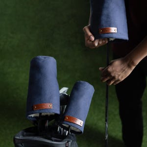 May include: A set of navy blue golf club head covers featuring a soft white interior lining and a rectangular brown leather patch on the front. Each patch is secured with two metallic rivets and displays embossed text on the surface.
