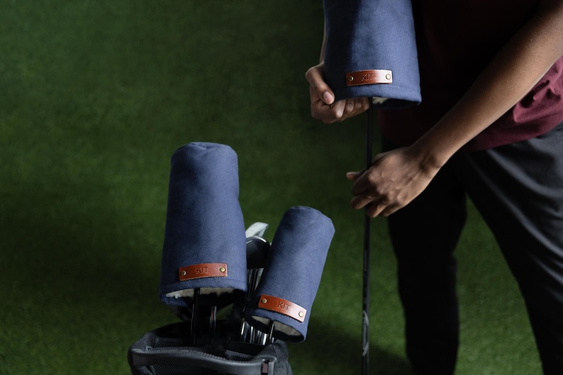 May include: A set of navy blue golf club head covers featuring a soft white interior lining and a rectangular brown leather patch on the front. Each patch is secured with two metallic rivets and displays embossed text on the surface.