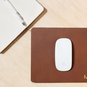 May include: A brown leather mousepad with a white mouse and a notebook with a pen. The mousepad has the name "MIA" in gold lettering in the bottom right corner. The notebook is open with blank white pages.