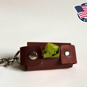 May include: A dark brown leather dog waste bag dispenser featuring a silver metal swivel clasp, two silver snap buttons, and a bright green plastic bag protruding from the center. A circular logo with the American flag and text Made in USA.