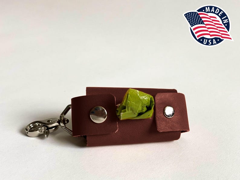May include: A dark brown leather dog waste bag dispenser featuring a silver metal swivel clasp, two silver snap buttons, and a bright green plastic bag protruding from the center. A circular logo with the American flag and text Made in USA.