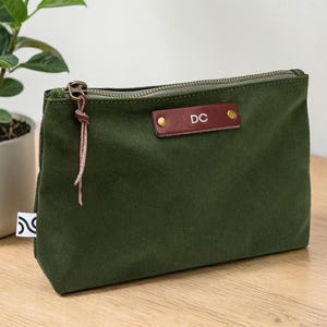 May include: An olive green canvas pouch with a brown leather tag displaying the initials "DC". The pouch has a metal zipper and a leather pull. A small plant in a white pot is in the background.