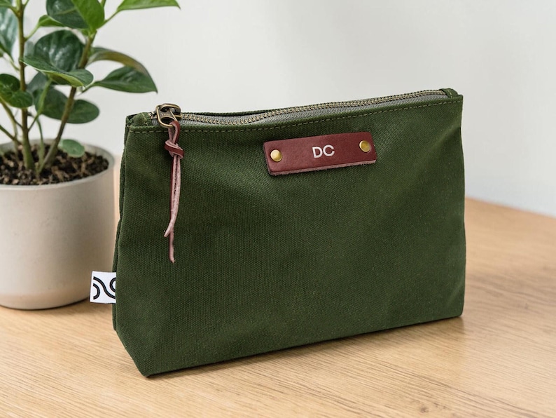 May include: An olive green canvas pouch with a brown leather tag displaying the initials "DC". The pouch has a metal zipper and a leather pull. A small plant in a white pot is in the background.