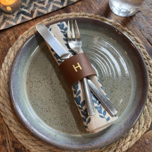 Puede incluir: Un juego de mesa con una servilleta beige y azul estampada, cubiertos y un servilletero de cuero marrón con la letra "H". El plato es gris moteado con un borde marrón, sobre un salvamanteles tejido.
