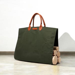 May include: An olive green canvas tote bag with brown leather handles and accents. The bag is filled with several cut logs. The handles are attached with brass rivets. The bag is designed for carrying firewood.