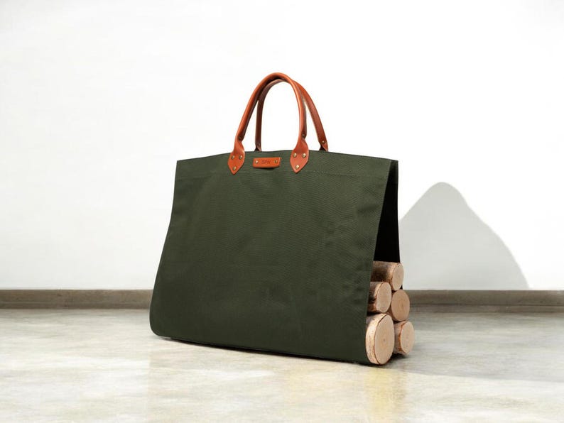May include: An olive green canvas tote bag with brown leather handles and accents. The bag is filled with several cut logs. The handles are attached with brass rivets. The bag is designed for carrying firewood.