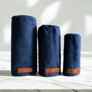 May include: Three navy blue golf club head covers of varying heights, each featuring a rectangular tan leather patch at the base. The leather patches are secured with two brass-colored rivets and are embossed with the initials JT in gold lettering.