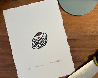 May include: A rectangular piece of off-white paper with deckled edges featuring a black ink block print of a brain with a small red heart on top. The text "RESILIENCE" is printed in black ink below the brain, accompanied by a handwritten signature.