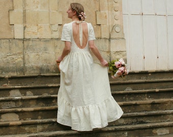 Pode incluir: Um vestido de noiva creme com costas baixas e mangas curtas. O vestido tem uma saia fluida e está sendo segurado por uma pessoa descendo degraus de pedra. Um buquê de flores cor-de-rosa é segurado na mão da pessoa.