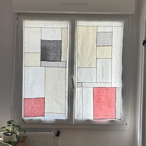 Pode incluir: Cortinas de janela com um design geométrico. As cortinas apresentam formas retangulares em branco, preto, vermelho e bege. Estão penduradas em frente a uma janela, com uma pequena mesa e uma planta visíveis em primeiro plano.