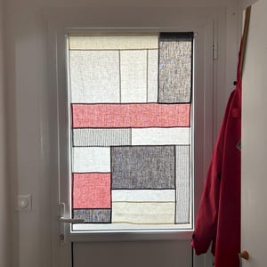 Pode incluir: Um tratamento de janela com padrão geométrico com um design de patchwork. O design incorpora retângulos em vermelho, preto, creme e branco. A janela está em uma moldura branca.