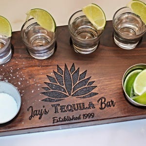 May include: A wooden tequila serving board with four shot glasses, each garnished with a lime wedge. A small bowl of salt and a bowl of lime wedges are also on the board. The board is engraved with "Jay's Tequila Bar Established 1999".