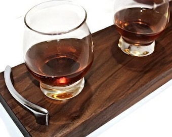 Puede incluir: Una bandeja de servir de madera con un asa de plata y cuatro vasos pequeños llenos de líquido ámbar. La bandeja es perfecta para una degustación de whisky.