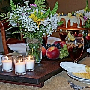 May include: A beautifully set table with a rustic wooden serving tray holding three lit votive candles. The table is adorned with fresh flowers, fruit, cake, and drinks, creating a warm and inviting atmosphere for a gathering.