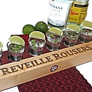 Puede incluir: Una bandeja de madera con seis vasos de chupito llenos de líquido transparente, adornados con rodajas de lima y cilantro. La bandeja tiene grabadas las palabras "REVEILLE ROUSERS" y el logotipo "ATM". Botellas de tequila y limas en el fondo.