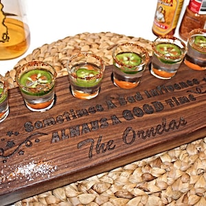 May include: A wooden shot glass tray with six shot glasses filled with clear liquid and green jalapeno peppers. The tray is engraved with the text "Sometimes a bad influence... ALWAYS a good time... The Ornelas".