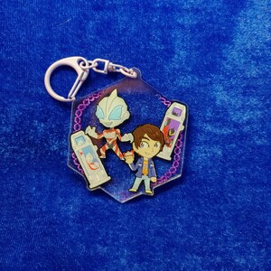 May include: A hexagonal acrylic keychain featuring a cartoon-style character with brown hair, a silver and red humanoid figure, and two rectangular devices with screen graphics. The design is bordered by a purple pattern on a clear acrylic base.