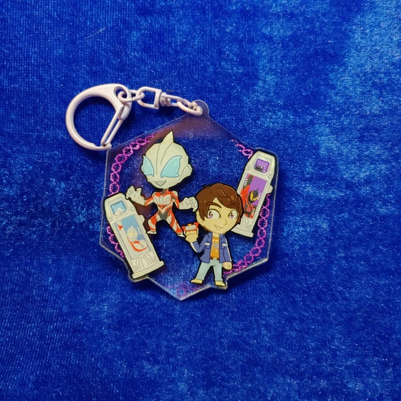 May include: A hexagonal acrylic keychain featuring a cartoon-style character with brown hair, a silver and red humanoid figure, and two rectangular devices with screen graphics. The design is bordered by a purple pattern on a clear acrylic base.