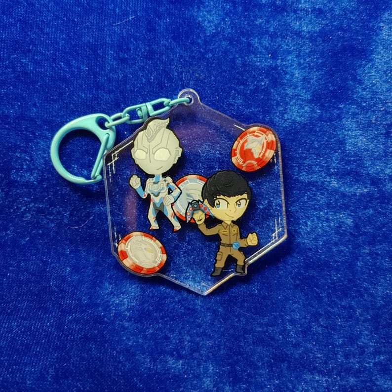 May include: A hexagonal clear acrylic keychain featuring a cartoon-style character in a silver and blue suit, a character in a brown uniform holding a small device, and three circular red and white tokens, attached to a light blue plastic clasp.