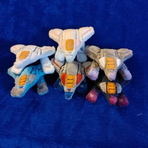 May include: A collection of plush toy airplanes in various colors, including blue, gray, and white. Each plane features orange and yellow accents, with details on the wings and body. The toys are arranged on a blue surface.