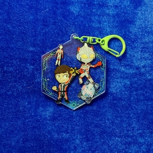 May include: A hexagonal acrylic keychain featuring a cartoon character with brown hair and a red and black suit, a stylized silver and red robot figure, and a small standing figure, all set against a dark blue background with green circuit lines.