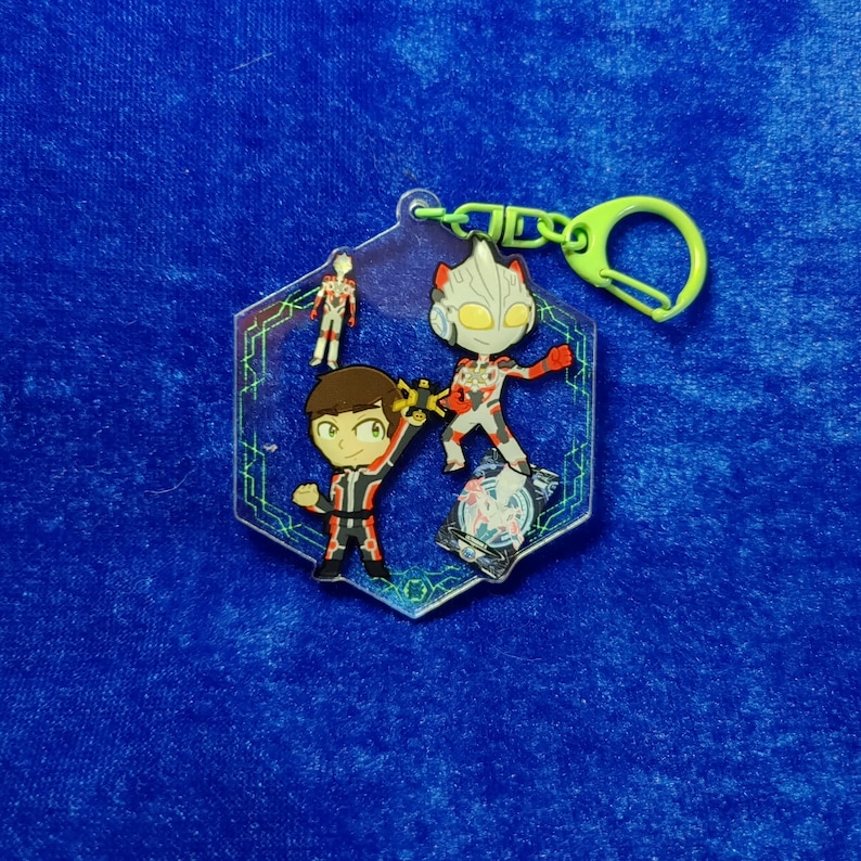May include: A hexagonal acrylic keychain featuring a cartoon character with brown hair and a red and black suit, a stylized silver and red robot figure, and a small standing figure, all set against a dark blue background with green circuit lines.