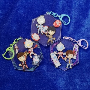 May include: Three hexagonal acrylic keychains featuring cartoon-style characters and small accessories. Each keychain includes a colored plastic clasp and displays various figures, including silver-masked heroes and human characters in uniform.