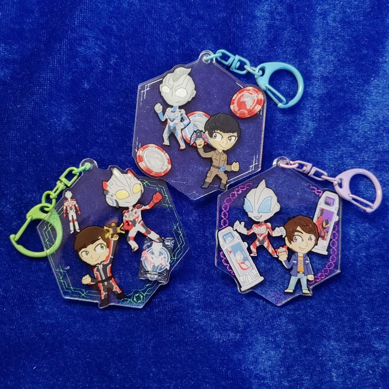 May include: Three hexagonal acrylic keychains featuring cartoon-style characters and small accessories. Each keychain includes a colored plastic clasp and displays various figures, including silver-masked heroes and human characters in uniform.
