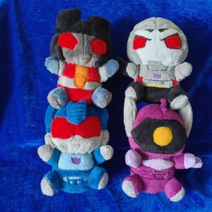 May include: Four plush toys in the image, each with a unique design. The toys are in the shape of characters, with red, blue, gray, and purple colors. The toys have arms and legs, and are stacked on top of each other.