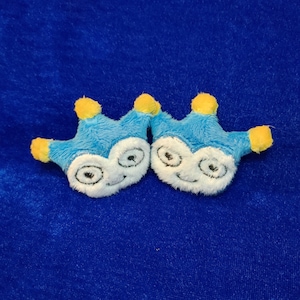 May include: Two plush blue and white toy crowns with yellow accents. Each crown has a smiling face with large eyes. The toys are on a blue background.