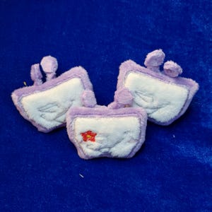 May include: Three plush, lavender-colored, square-shaped items with white centers and small, rounded, lavender protrusions. One item has a small red flower embroidered on it. The items are arranged on a blue surface.
