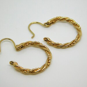 May include: A pair of gold-colored hoop earrings. The earrings have a twisted rope design with small, evenly spaced beads. The hooks are curved and open. The earrings are on a white background.