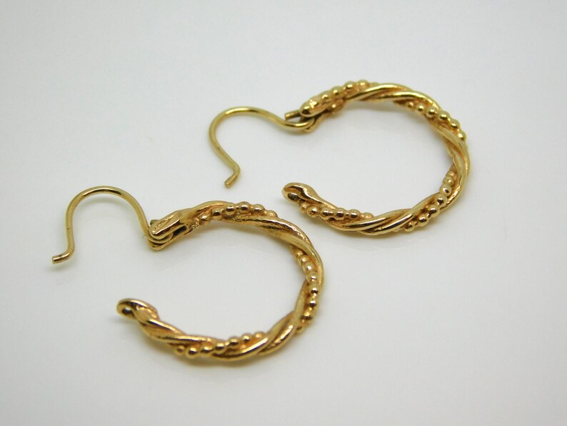 May include: A pair of gold-colored hoop earrings. The earrings have a twisted rope design with small, evenly spaced beads. The hooks are curved and open. The earrings are on a white background.