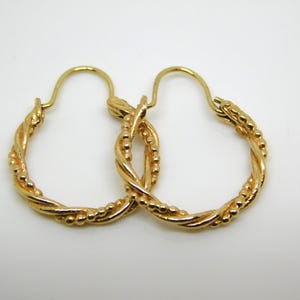 May include: A pair of gold-colored hoop earrings. The earrings have a twisted rope design with small beaded accents. The hoops are displayed on a white background, showcasing the jewelry's details and craftsmanship.
