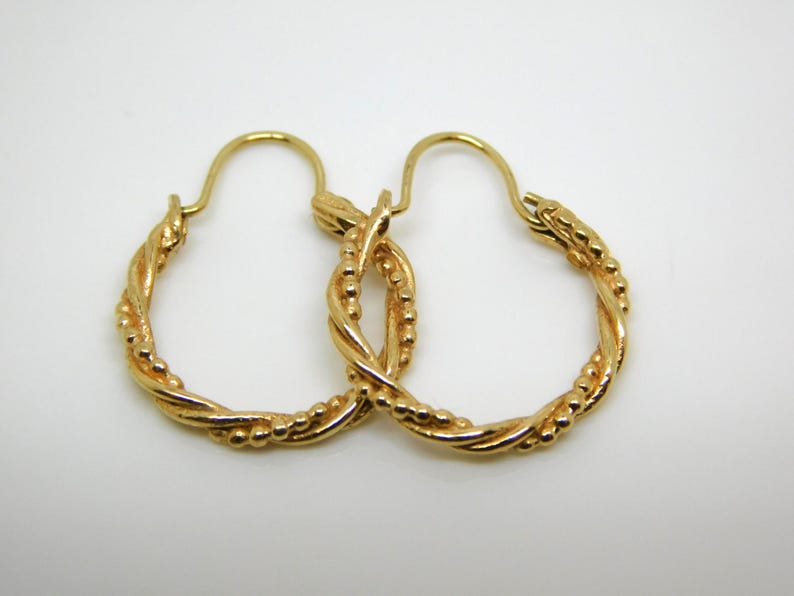 May include: A pair of gold-colored hoop earrings. The earrings have a twisted rope design with small beaded accents. The hoops are displayed on a white background, showcasing the jewelry's details and craftsmanship.