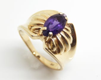 May include: A gold-colored ring featuring a marquise-cut, deep purple gemstone. The ring has a decorative, fan-like design around the gemstone, with a smooth, polished band. The ring is set against a white background.