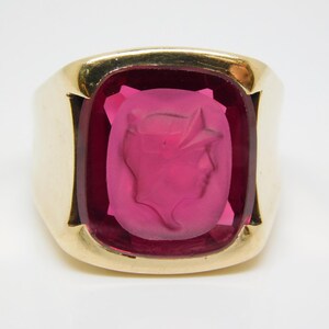 May include: A gold ring with a large, rectangular, pink gemstone. The gemstone features a carved profile of a person. The ring has a thick band and a classic design, suitable for a variety of occasions.