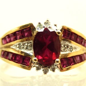 May include: A gold ring featuring a large oval-cut red gemstone at the center, surrounded by small clear stones. The band is accented with rows of square-cut red stones set in gold channels, with additional clear stones embedded in the metal setting.
