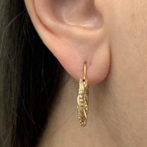 May include: Close-up of a gold-colored earring. The earring is a small hoop with a textured, braided design. The earring is attached to an ear. The earring is made of a shiny, metallic material.