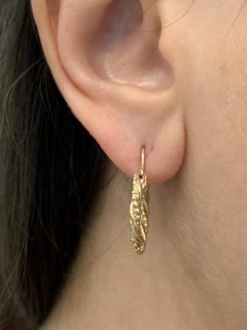 May include: Close-up of a gold-colored earring. The earring is a small hoop with a textured, braided design. The earring is attached to an ear. The earring is made of a shiny, metallic material.