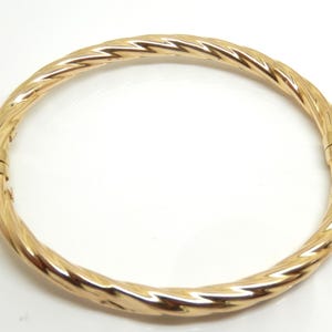 May include: A polished gold-tone bangle bracelet featuring a continuous twisted rope design. The circular bracelet has a smooth, reflective metallic surface and includes a small, integrated hinged clasp mechanism on one side for opening and closing.
