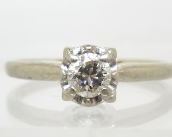 May include: A silver ring with a single round diamond set in a prong setting. The diamond is surrounded by a decorative border.